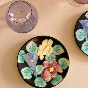 May include: A black ceramic plate featuring a floral design. The flowers are painted in shades of yellow, blue, green, pink, and gold. The plate is ideal for serving snacks or desserts.