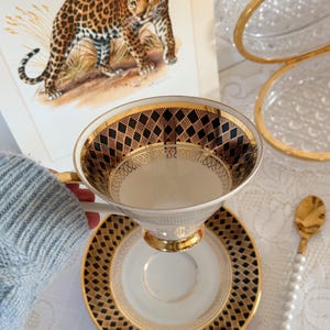 Peut inclure: Service à thé vintage avec une tasse et une soucoupe à bordure dorée et motif géométrique noir. La tasse est tenue au-dessus de la soucoupe, mettant en valeur le design complexe. Une cuillère dorée avec un manche en perles est à proximité. Illustration de léopard en arrière-plan.