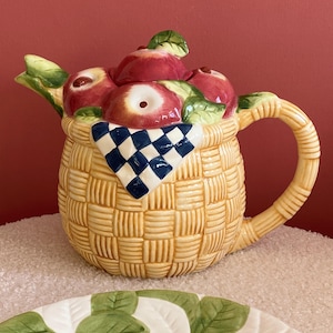 May include: A ceramic teapot shaped like a basket overflowing with red apples. The basket has a blue and white checkered cloth on top. The teapot has a handle and a spout.