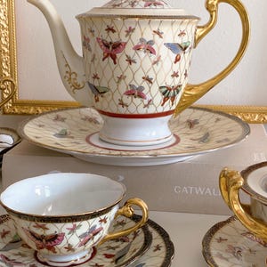 Può includere: Un set da tè vintage con un design a farfalla. La teiera, le tazze e i piattini sono bianchi con finiture dorate e presentano farfalle colorate su uno sfondo crema. Il set ha un'estetica delicata e femminile.