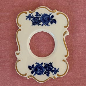 Könnte beinhalten: Weiße Porzellan-Lichtschalterabdeckung mit dekorativem Blumendesign in Blau und Gold. Die Abdeckung hat eine zentrale kreisförmige Öffnung und ist mit blauen Rosenbouquets und goldenen Zierleisten verziert.