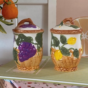 Peut inclure: Deux pots en céramique avec couvercles, présentant des motifs de fruits. L'un a un motif de raisin violet, l'autre des citrons. Les deux pots ont un motif de tissage de panier et des poignées marron. Les pots sont sur une surface rayée.