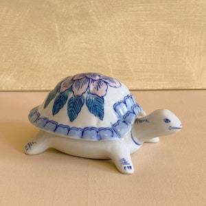 Peut inclure: Une boîte à bijoux en forme de tortue en céramique blanche avec un motif floral bleu et rose sur le dessus. La tortue est posée sur une surface beige clair.