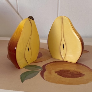 May include: Two pear-shaped decorative objects, each cut in half, with a glossy finish. The pears transition from yellow to reddish-brown, with a dark brown stem. They sit on a surface with a peach and leaf design.