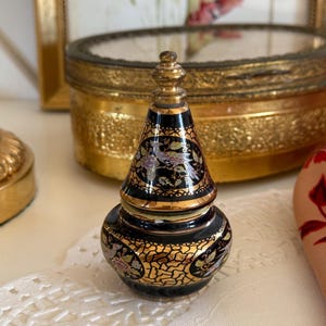 Può includere: Un piccolo vasetto decorativo in ceramica con coperchio conico, caratterizzato da una finitura nera lucida decorata con intricati motivi in filigrana dorata e disegni floreali colorati. L'oggetto presenta un pomello dorato a punta.