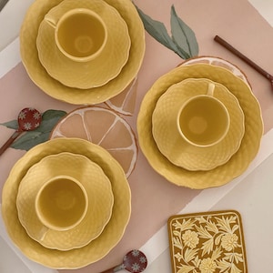 Puede incluir: Un juego de tazas y platillos de cerámica amarilla con un patrón texturizado. Las tazas están dispuestas sobre un mantel individual con estampado rosa y blanco con un diseño floral. También hay cucharas de madera con extremos decorativos y un posavasos floral.