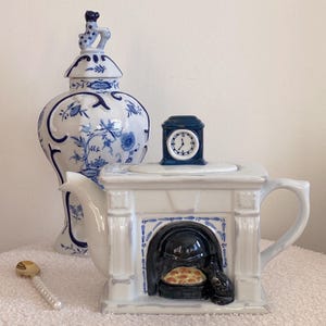 May include: White ceramic teapot shaped like a fireplace with a clock on top. The fireplace has a black and blue interior with a small black cat sitting in front of it.