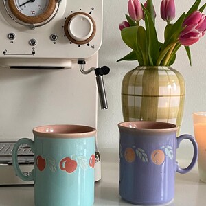 May include: Two ceramic mugs with a pink rim and a fruit design. The mug on the left is light blue with a cherry design. The mug on the right is purple with an orange design.