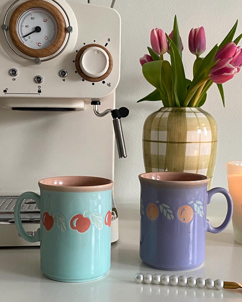 May include: Two ceramic mugs with a pink rim and a fruit design. The mug on the left is light blue with a cherry design. The mug on the right is purple with an orange design.