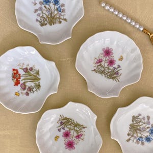 May include: Five white ceramic seashell-shaped dishes with floral designs. Each dish features a different arrangement of flowers in shades of pink, blue, yellow, and red.