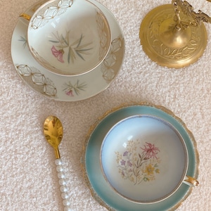 Puede incluir: Dos tazas de té y platillos con motivos florales. Las tazas de té son blancas con ribetes dorados y los platillos son blancos con ribetes dorados y azules. También se muestra una cuchara dorada con mango de perla.