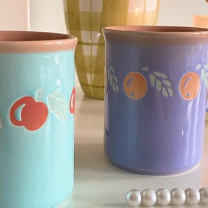 May include: Two ceramic mugs with a pastel blue and purple glaze. The mugs have a pink rim and feature a white and orange fruit design. The mugs are sitting on a white surface with a string of white pearls.