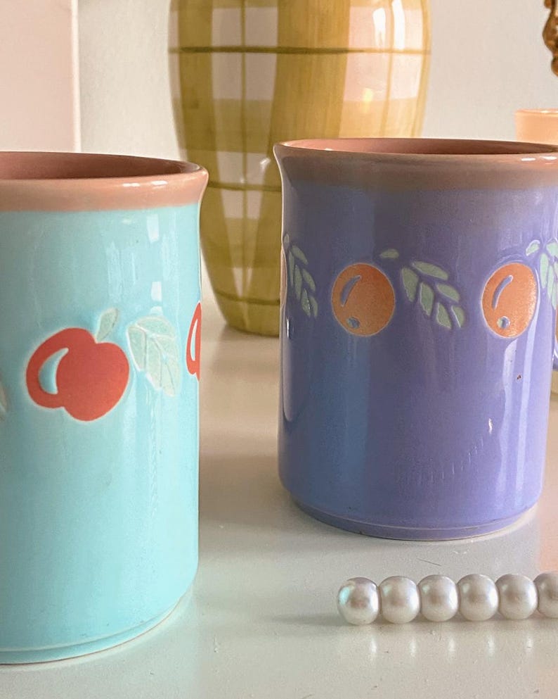 May include: Two ceramic mugs with a pastel blue and purple glaze. The mugs have a pink rim and feature a white and orange fruit design. The mugs are sitting on a white surface with a string of white pearls.
