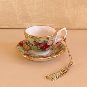 Puede incluir: Un adorno de taza de té y platillo en miniatura con un patrón floral en tonos verdes, rojos y amarillos. El adorno tiene un borde dorado y una borla dorada.