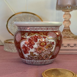 Könnte beinhalten: Dekorative Keramiktopf mit weißem Rand und einem rot-goldenen Blumendesign. Der Topf weist ein detailliertes Muster aus Blumen und Blättern auf. Der Topf steht auf einer rosa Oberfläche. Das Design des Topfes erinnert an orientalische Keramik.
