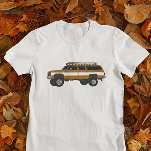 May include: White t-shirt with a brown and white illustration of a vintage SUV with a roof rack.