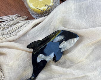 May include: A black and white orca hair clip made of plastic. The orca is in a swimming position with its tail fin extended.