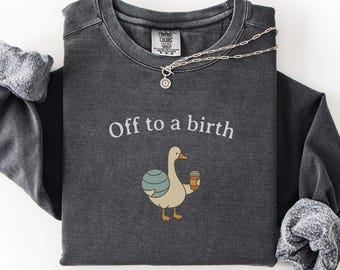 May include: Dark grey long-sleeved sweatshirt featuring the text "Off to a birth" and a cartoon goose holding a coffee cup. The goose has a blue and green patterned egg on its back. A silver necklace with a small pendant is draped on the sweatshirt.