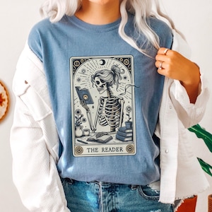 May include: A blue t-shirt with a graphic of a skeleton reading a book. The graphic is in a tarot card style with the text "THE READER" at the bottom.