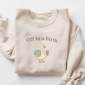 May include: Cream-colored sweatshirt with the text "Off to a birth." The graphic features a cartoon duck holding a coffee cup and a blue and white ball. The sleeves are rolled up.