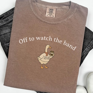 May include: A brown Comfort Colors t-shirt with the text "Off to watch the band" and a cartoon duck holding a coffee cup. The duck has a scarf and a "Here for Halftime" sign. The shirt is paired with black jeans and white sneakers.
