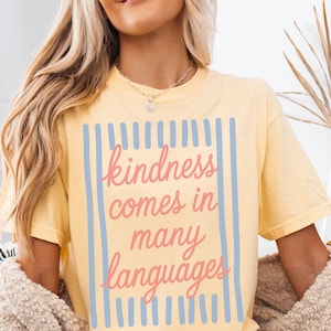 May include: A light yellow t-shirt with the text "kindness comes in many languages" in pink script and blue vertical lines. The shirt is made of a soft material and is a comfortable fit.