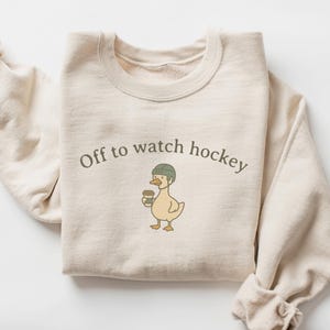 May include: A cream-colored crewneck sweatshirt featuring a graphic of a duck wearing a green hockey helmet and holding a coffee cup. The text "Off to watch hockey" is printed in dark gray lettering above the duck illustration on the chest.
