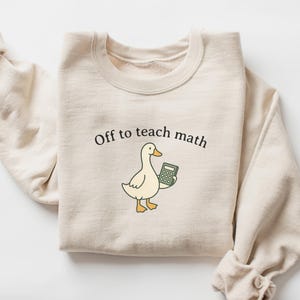 May include: Cream-colored sweatshirt with the text "Off to teach math." A cartoon duck holding a calculator is printed below the text. The sweatshirt has long sleeves and a crew neck.