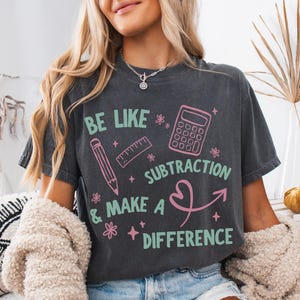 May include: A charcoal gray short-sleeve t-shirt featuring a graphic print in light green and pink. The design includes a pencil, ruler, and calculator with the text: BE LIKE SUBTRACTION & MAKE A DIFFERENCE, accented by small star and flower icons.