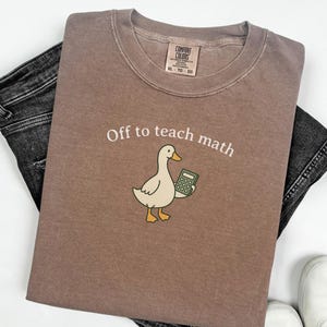 May include: A brown t-shirt with the text "Off to teach math" and a cartoon duck holding a calculator. The shirt is paired with black jeans and white sneakers. The shirt has a Comfort Colors tag.