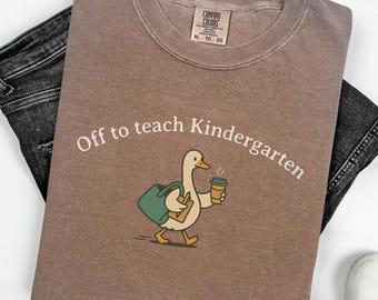 May include: A brown Comfort Colors t-shirt with the text "Off to teach Kindergarten" and a cartoon duck carrying a bag and coffee cup. The shirt is paired with black jeans and white sneakers.