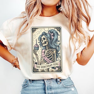 May include: A white t-shirt featuring a graphic of a skeleton holding a cupcake and a sash that reads "It's my 30th" and "The Birthday Girl".