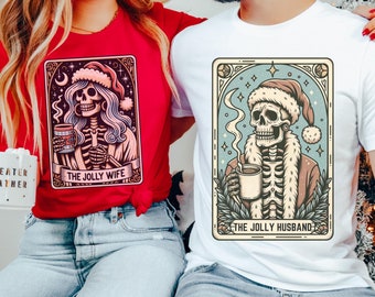 May include: A couple wearing matching Christmas themed t-shirts. The woman's shirt is red and features a skeleton wearing a Santa hat and holding a mug. The text on her shirt reads "The Jolly Wife". The man's shirt is white and features a skeleton wearing a Santa hat and holding a mug. The text on his shirt reads "The Jolly Husband".