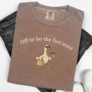 May include: A brown Comfort Colors t-shirt with the phrase "Off to be the fun aunt" and a cartoon duck wearing sunglasses and carrying a coffee cup and shopping bag. The shirt is laid on top of black denim jeans and white sneakers.