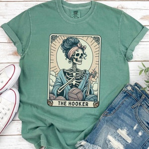 May include: A green t-shirt with a graphic of a skeleton woman crocheting with yarn balls and the text "THE HOOKER".