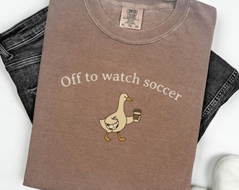 Może przedstawiać: Brązowa koszulka Comfort Colors z napisem "Off to watch soccer" i rysunkiem kaczki trzymającej kubek z kawą. Koszulka jest zestawiona z czarnymi dżinsami i białymi trampkami.