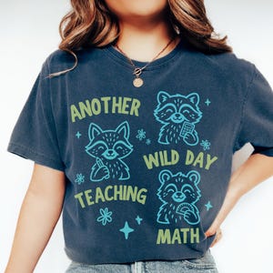 May include: A dark blue short-sleeve t-shirt featuring three raccoon illustrations in light blue and green. The text reads "ANOTHER WILD DAY TEACHING MATH" with small star and flower accents. The shirt has a crew neckline and a relaxed fit.