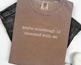 May include: A brown t-shirt with the text "maybe sourdough is obsessed with me" in white. The shirt is folded over a pair of black jeans. White sneakers are visible in the bottom right corner. The shirt has a Comfort Colors tag.