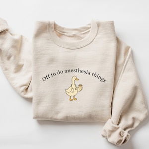 Puede incluir: Sudadera color crema con la frase "Off to do anesthesia things" impresa en negro. Un pato de dibujos animados con estetoscopio y taza de caf&eacute; est&aacute; centrado debajo del texto. La sudadera tiene cuello redondo y mangas largas.