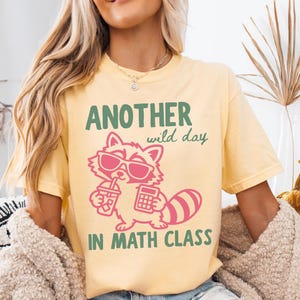 May include: A butter yellow t-shirt featuring a graphic of a raccoon wearing pink sunglasses, holding a drink and a calculator. The text reads "ANOTHER wild day IN MATH CLASS" in green lettering. The design is centered on the front of the shirt.