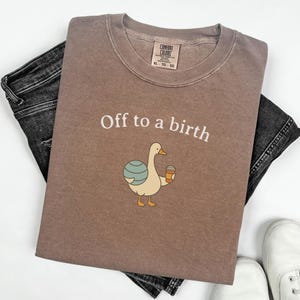 Może przedstawiać: Brązowa koszulka z napisem "Off to a birth" i kreskówkową kaczką trzymającą kubek z kawą. Koszulka jest ułożona na czarnych dżinsach, widoczne są białe trampki.