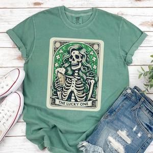 May include: Sage green t-shirt featuring a graphic of a skeleton holding a beer, with green hair and a decorative border. The text "THE LUCKY ONE" is below the image. The shirt is paired with denim shorts and white sneakers.
