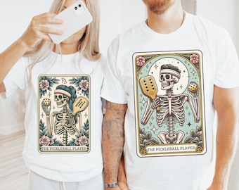 May include: Two white t-shirts with a skeleton playing pickleball graphic. The graphic is in a vintage style with a blue background and floral accents. The text on the graphic reads "The Pickleball Player".