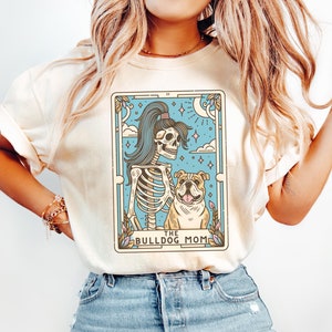 May include: A white t-shirt featuring a graphic of a skeleton holding a bulldog. The graphic is in a tarot card style with the text "The Bulldog Mom".