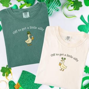 May include: Two t-shirts, one green and one cream, with the phrase "Off to get a little silly" and a cartoon duck wearing a hat and holding a beer. The shirts are surrounded by St. Patrick's Day decorations.