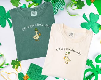 May include: Two t-shirts, one green and one cream, with the phrase "Off to get a little silly" and a cartoon duck wearing a hat and holding a beer. The shirts are surrounded by St. Patrick's Day decorations.