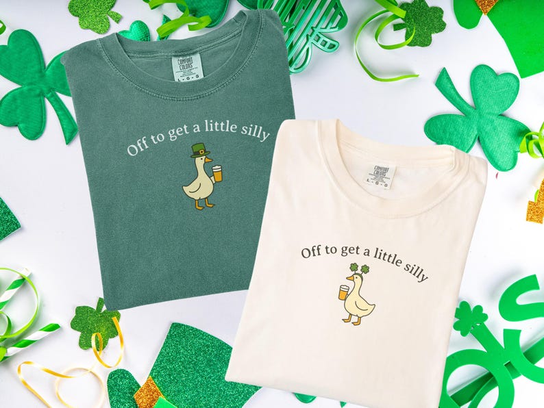 May include: Two t-shirts, one green and one cream, with the phrase "Off to get a little silly" and a cartoon duck wearing a hat and holding a beer. The shirts are surrounded by St. Patrick's Day decorations.