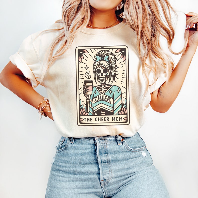Pu&ograve; includere: Una maglietta beige con una grafica di uno scheletro che indossa un costume da cheerleader blu e bianco e tiene una tazza di caff&egrave;. La grafica &egrave; in stile carta dei tarocchi e dice "The Cheer Mom" in basso.
