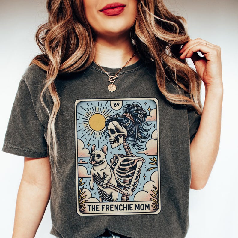 May include: A dark gray t-shirt with a graphic of a skeleton holding a French bulldog. The graphic is in the style of a tarot card and says "The Frenchie Mom" at the bottom.