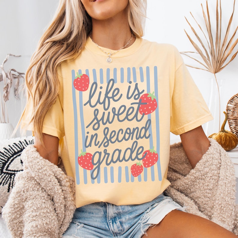 K&ouml;nnte beinhalten: Hellgelbes T-Shirt mit dem Schriftzug "Life is sweet in second grade" in blauer Schrift, umrahmt von blauen vertikalen Streifen und roten Erdbeer-Illustrationen. Ein l&auml;ssiges Kurzarm-Oberteil.